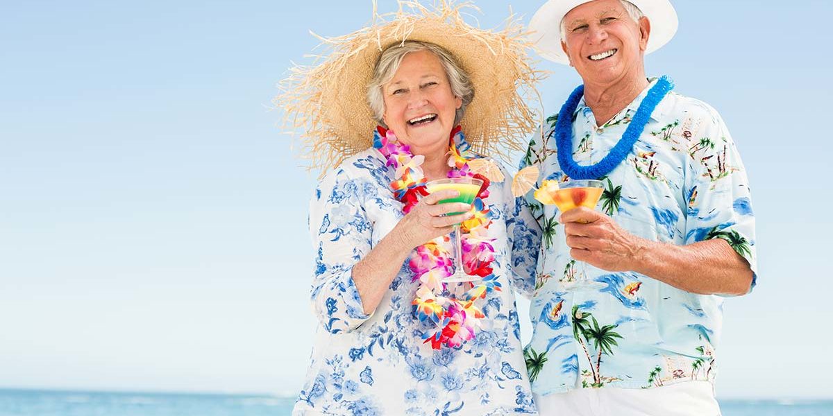 Wohnen im Ausland Seniorenehepaar mit Blumenketten, Hawaiihemden und Sonnenhüten genießt Cocktails am Strand | Wohnen im Ausland