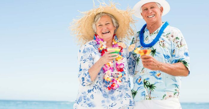 Wohnen im Ausland Seniorenehepaar mit Blumenketten, Hawaiihemden und Sonnenhüten genießt Cocktails am Strand | Wohnen im Ausland