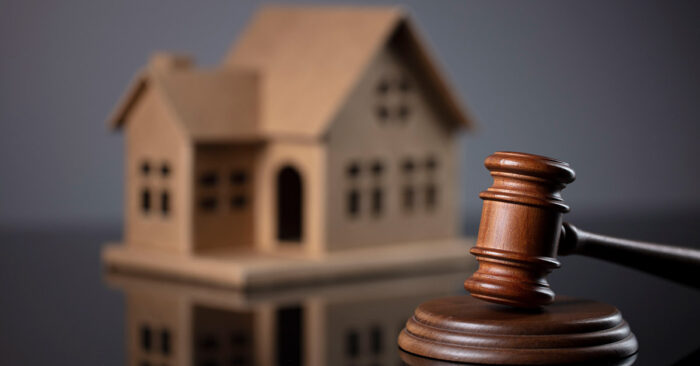 Ein Gavel, oder Richterhammer, aus Holz liegt auf seinem Resonanzblock aus Holz auf einem glänzenden schwarzen Tisch und im Hintergrund steht ein Hausmodell aus Holz | Lebenslanges Wohnrecht