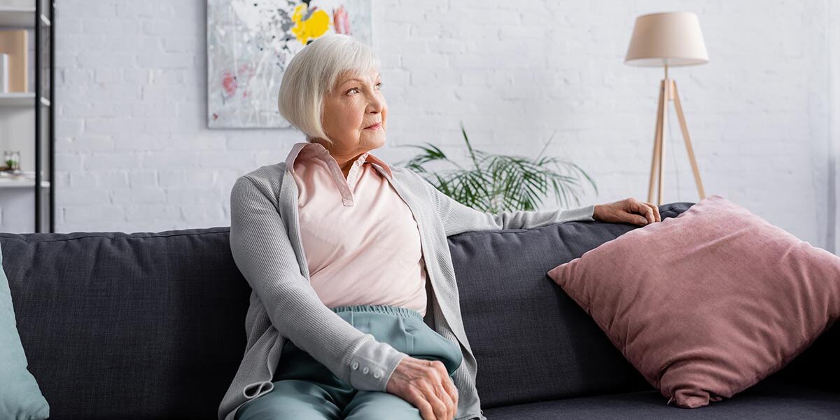 Wohnen im Alter Eine Seniorin sitzt auf ihrem Sofa in ihrem Wohnzimmer, im Hintergrund hängt ein modernes Gemälde, daneben stehen eine Topfpflanze, eine Stehlampe und ein Regal | Wohnen im Alter