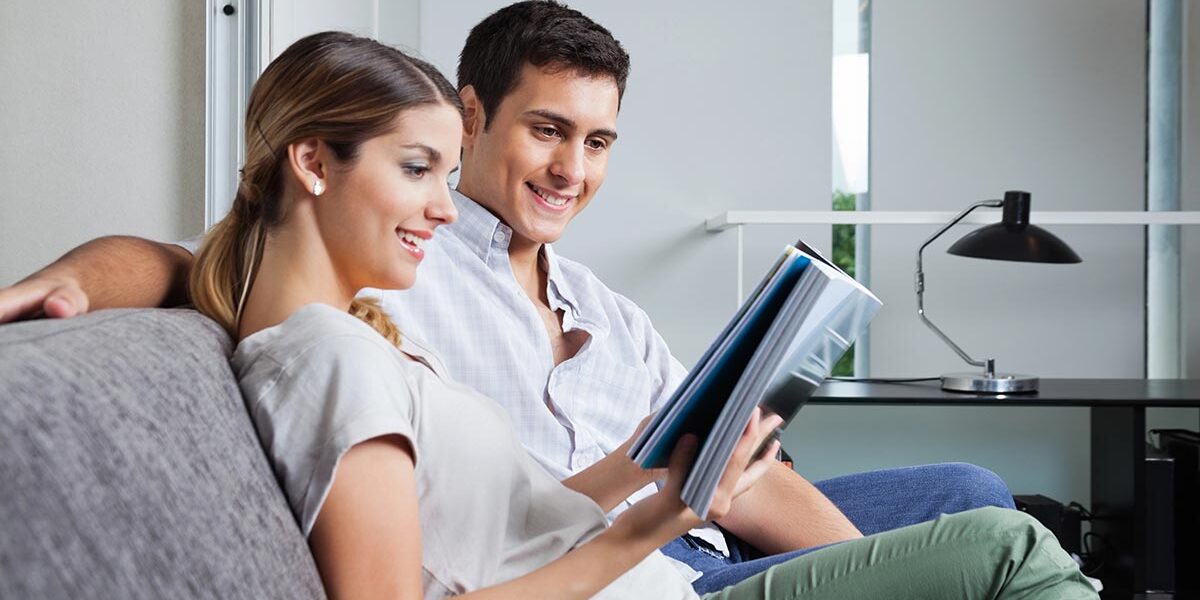 Ein junges Paar durchblättert auf dem Sofa in seiner Mietwohnung ein Exposé für eine Immobilie, die sie vielleicht kaufen möchten | Immobilienverkauf