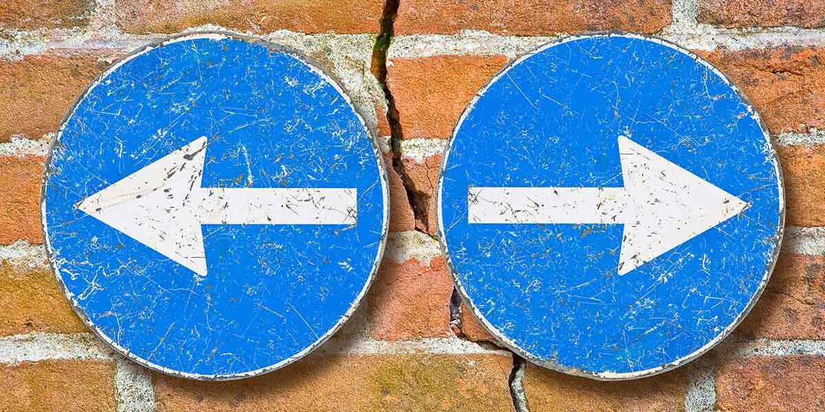 Scheidungsimmobilie Durch eine Backsteinmauer, als Symbol eines Hauses, verläuft fast senkrecht ein Riss, rechts und links davon befinden sich zwei blaue Schilder mit weißen Pfeilen, die jeweils nach rechts und links, also vom Riss weg, zeigen, als Symbol für das Scheidungspaar, dass getrennte Wege geht | Scheidungsimmobilie