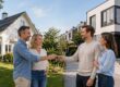 Vier Erwachsene stehen zwischen einem Einfamilienhaus und einer modernen Stadtvilla und übergeben sich symbolisch einen Schlüssel als Zeichen für einen Immobilientausch. | Immobilientausch