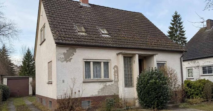 Ein älteres Einfamilienhaus mit verwitterter Fassade und sanierungsbedürftigem Dach steht in einer Wohnstraße und symbolisiert die Entscheidung zwischen Modernisierung und Verkauf. I Sanierungspflicht beim Verkaufen