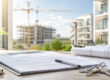 Kaufunterlagen, Baupläne und Schlüssel auf einem Schreibtisch vor moderner Neubauarchitektur – symbolisch für den Kauf einer Immobilie vom Bauträger. I Kauf vom Bauträger