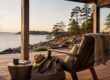 Sonnige Terrasse einer Ferienimmobilie am Wasser mit Sessel, Decke, Reisetasche und Blick auf eine bewaldete Küste I Ferienimmobilie