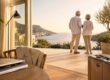 Älteres Paar blickt von der Terrasse seines Hauses auf eine sonnige Küstenlandschaft – Sinnbild für einen Lebensabend im Ausland.
